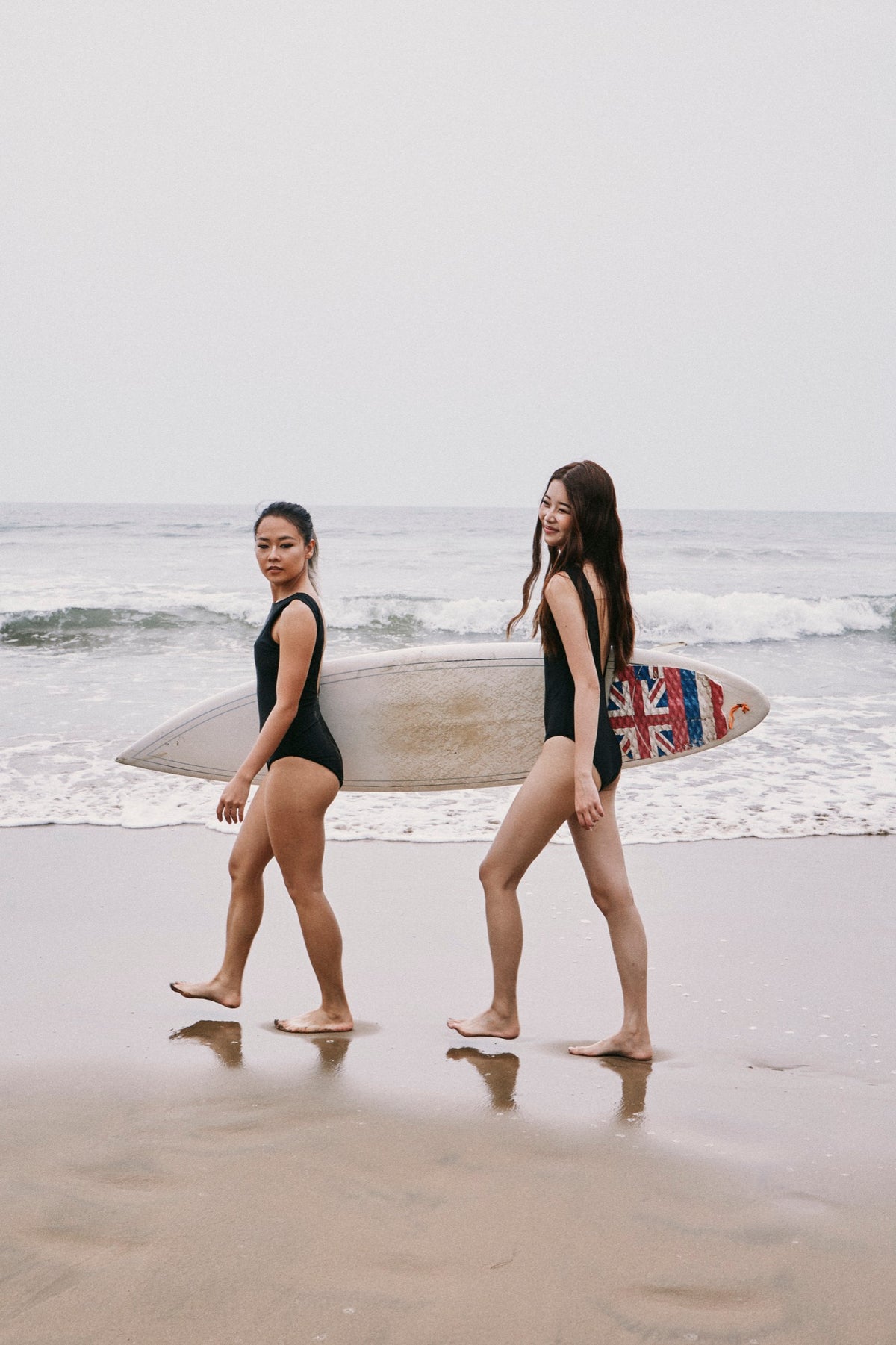 Two women in black B.Peachy swimsuits carrying a surfboard along the beach